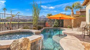 A backyard with a swimming pool, a hot tub, and a patio area with chairs and an umbrella.