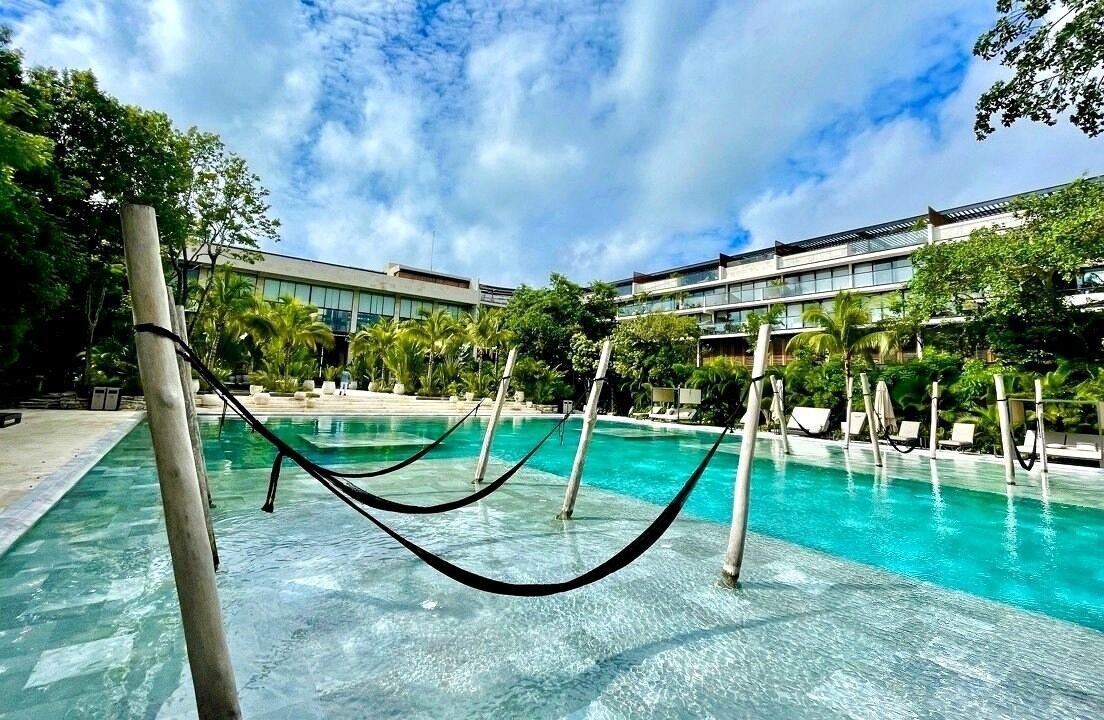 A pool area with clear water, surrounded by lounge chairs and palm trees, with a building in the background.