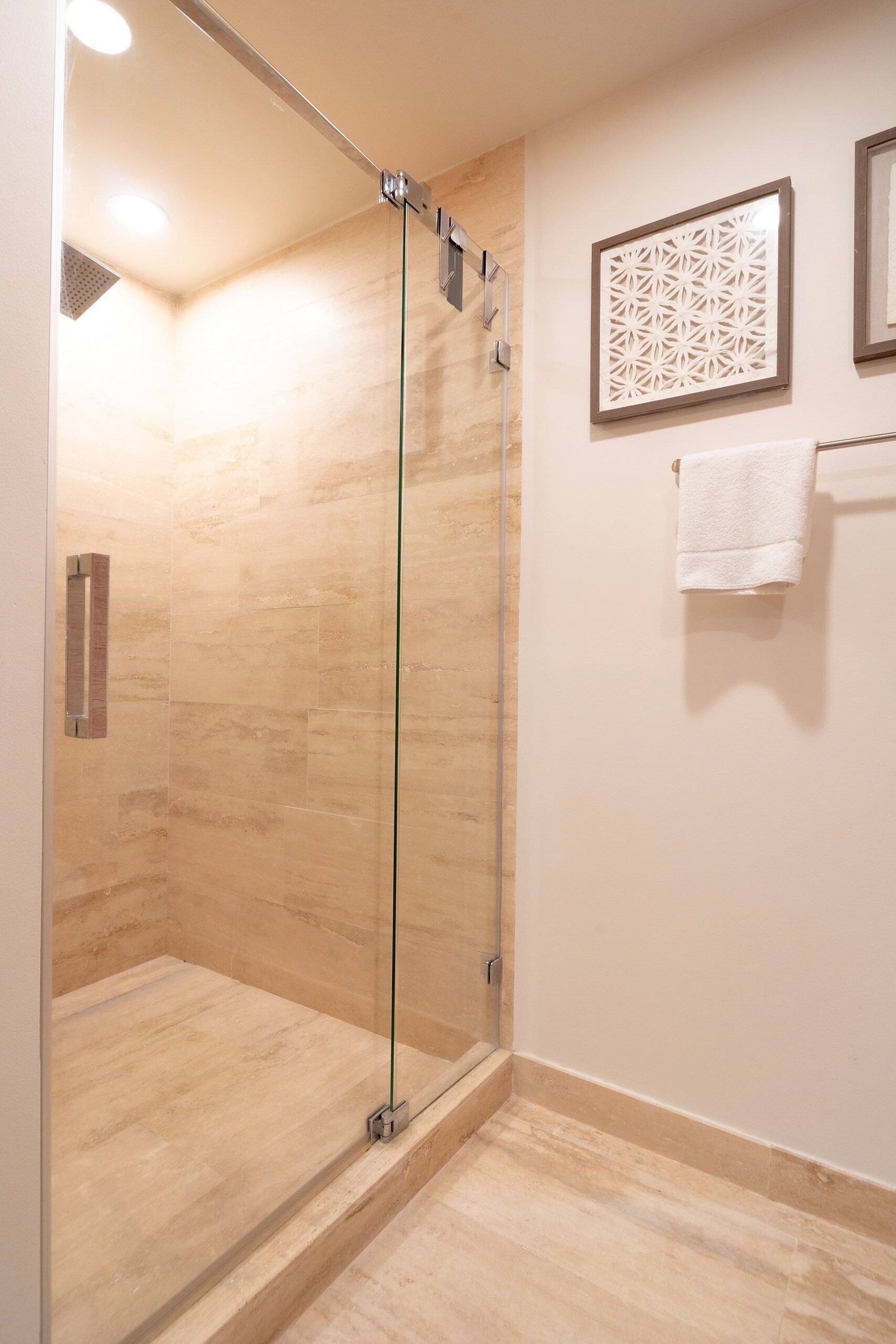 A glass-enclosed shower with a tiled floor, a towel rack with a white towel, and a decorative wall panel.