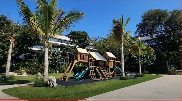 Un parque infantil con toboganes y estructuras para escalar, rodeado de palmeras y un césped cuidado.