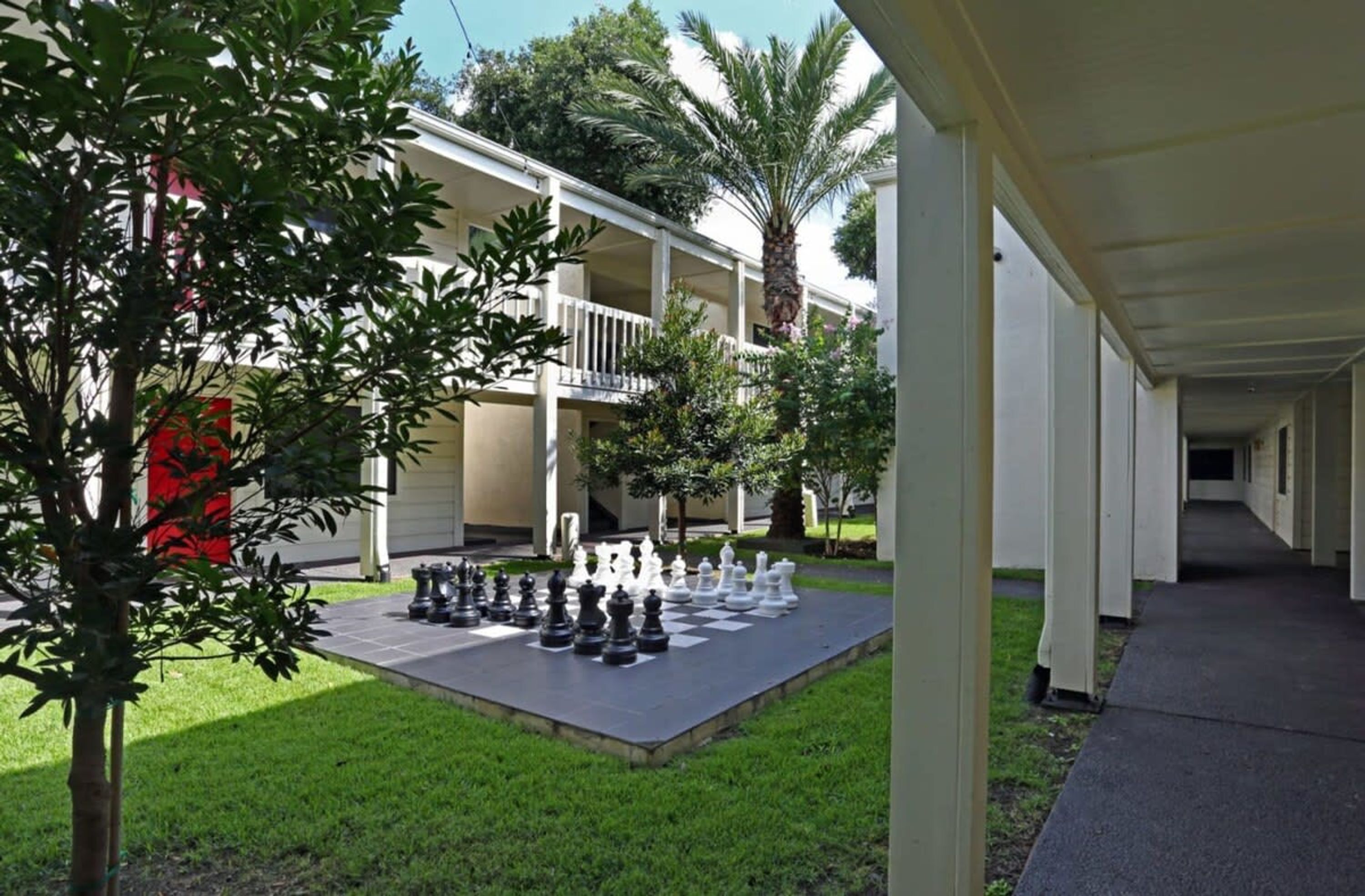 A courtyard with a chessboard set up, surrounded by a building with a covered walkway and palm trees.