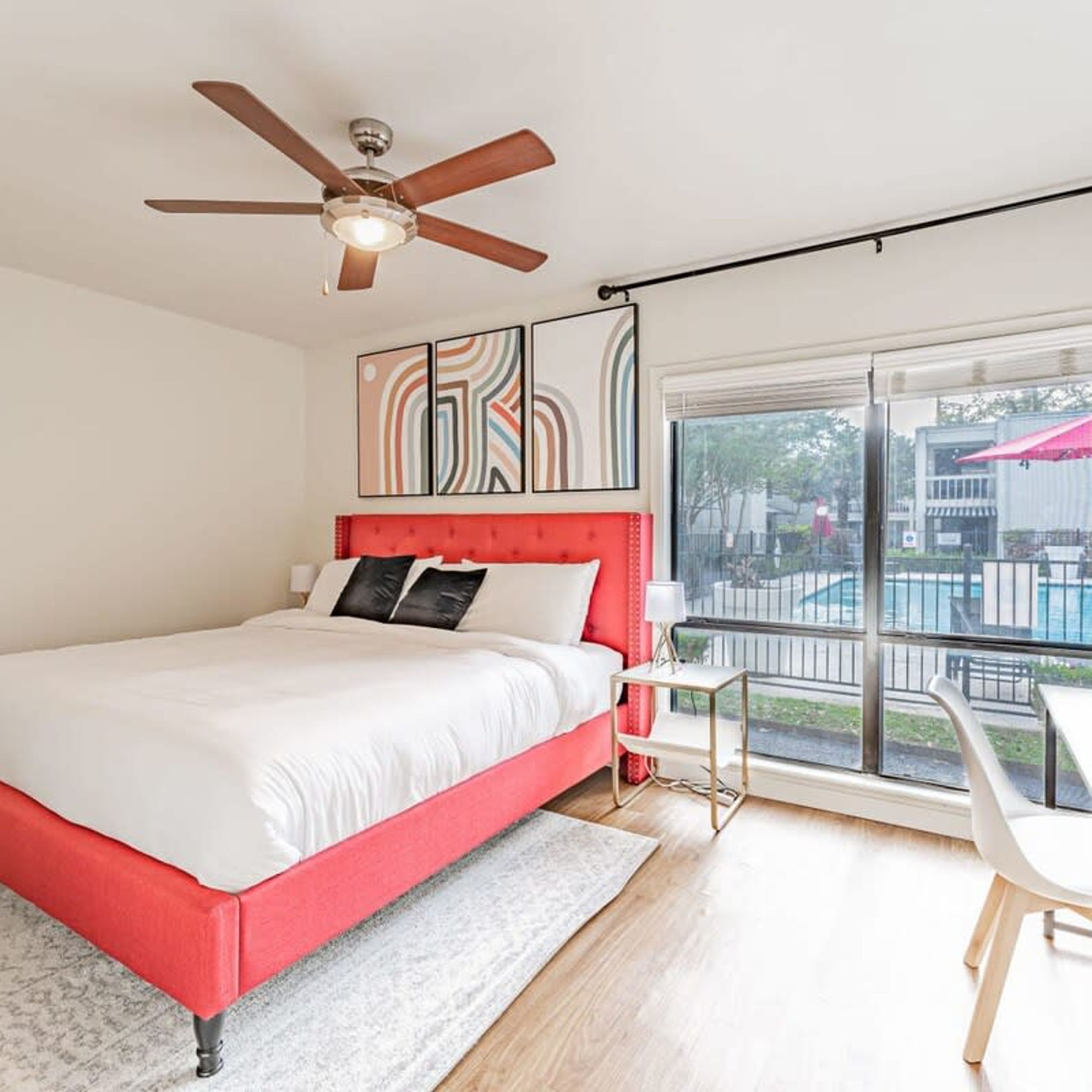 A bedroom with a red bed, a ceiling fan, a large window with a view of a pool, and a small table by the window.