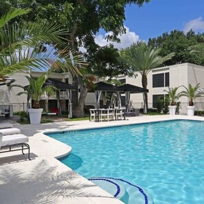 A swimming pool with lounge chairs and umbrellas, surrounded by trees and a building with a flat roof. - Chic 2BR Near Triangle With King Beds Fast Wifi (Austin)