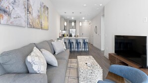 Una sala de estar moderna con un sofá gris, una mesa de centro y un televisor. Se ve una cocina al fondo con taburetes de bar y un refrigerador.