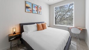 Un dormitorio con cama, una mesita de noche con lámpara, ventilador de techo, ventana con persianas y una silla.