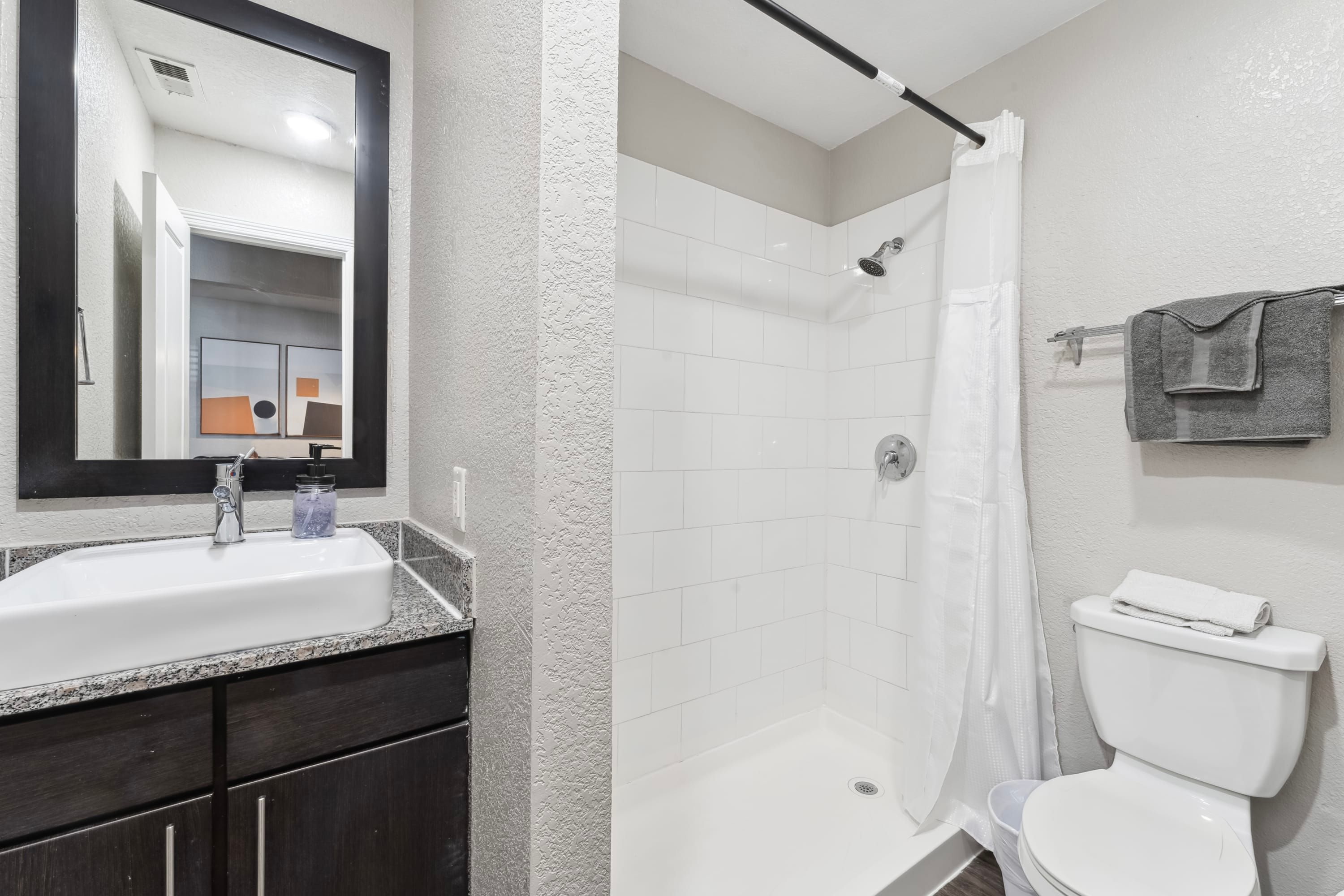 Een badkamer met een witte wastafel, donkere kastjes, een douche met een witte gordijn en een toilet met een handdoek erop.