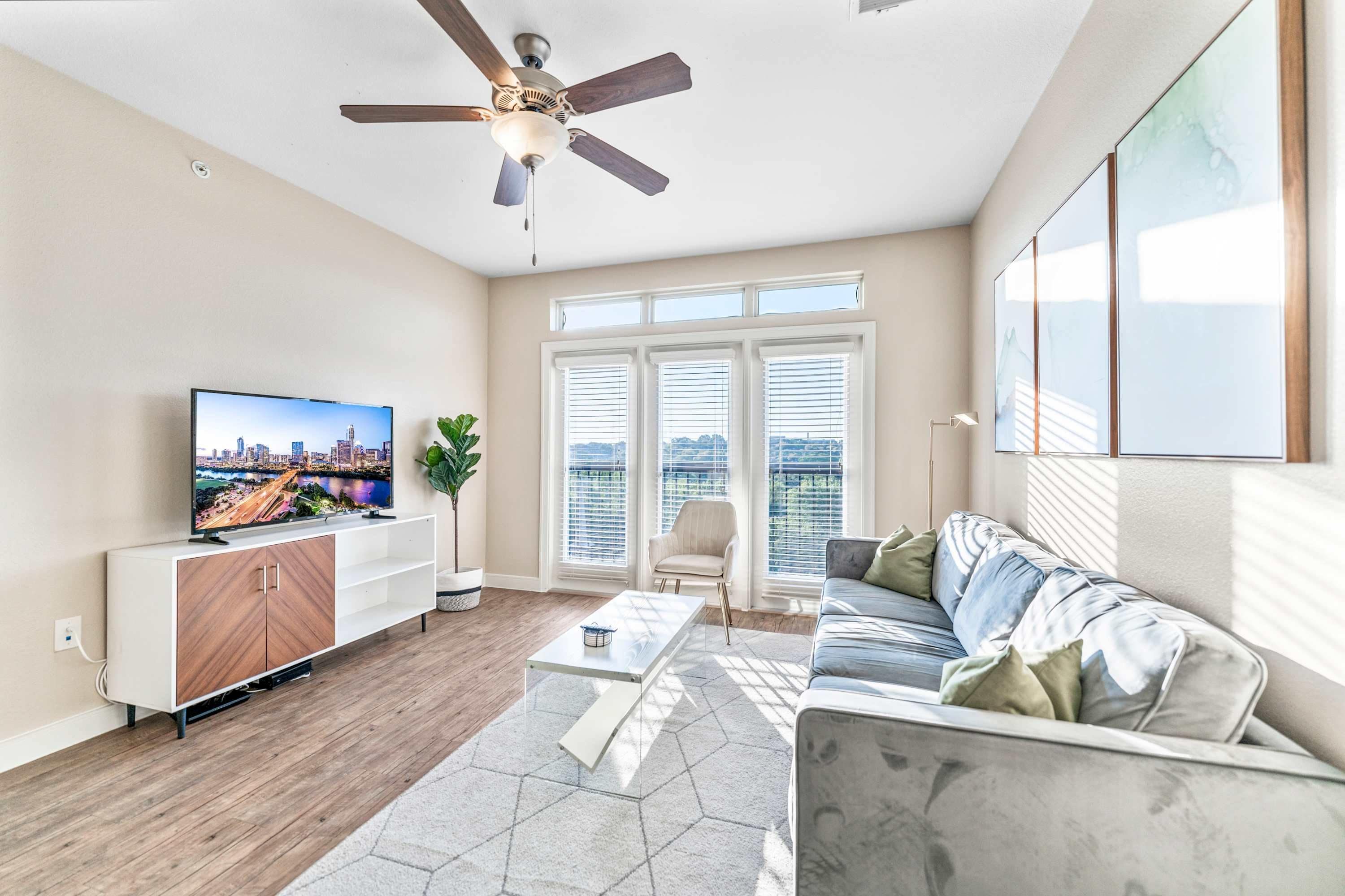 A modern living room with a flat-screen TV, a ceiling fan, a sofa, and a large window with blinds.