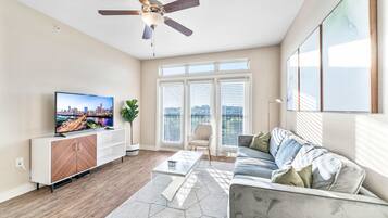 A modern living room with a flat-screen TV, a ceiling fan, a sofa, and a large window with blinds.
