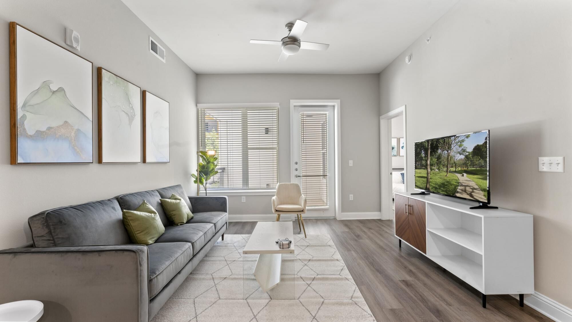 A modern living room with a gray sofa, a coffee table, and a TV on a white stand. There are three framed artworks on the wall, and a ceiling fan is visible.