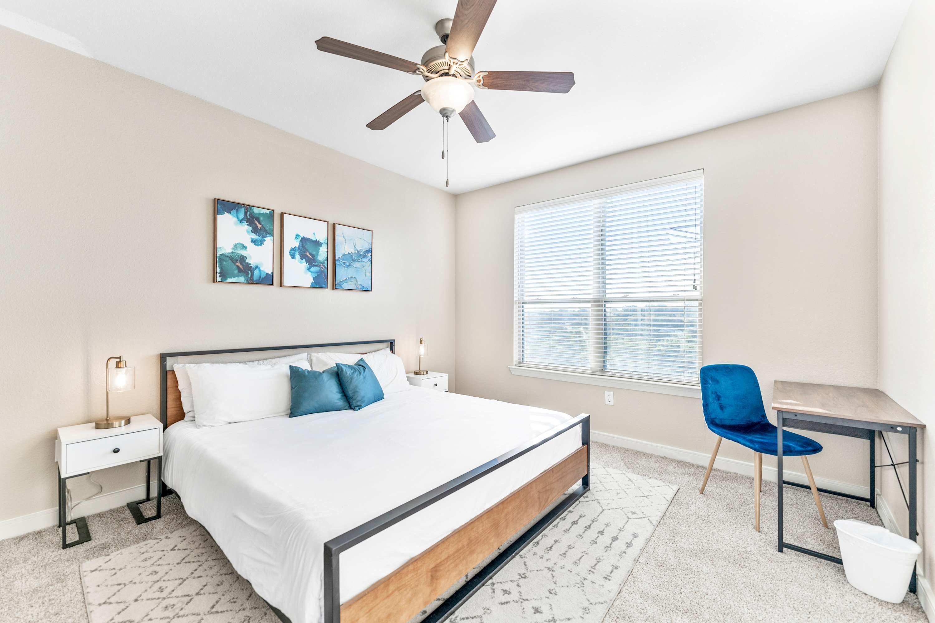 Une chambre à coucher comprenant un lit, des tables de chevet, un bureau, une chaise, un ventilateur de plafond et une fenêtre avec des stores.