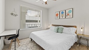 A bedroom with a bed, a desk, a chair, a ceiling fan, and a window with blinds.