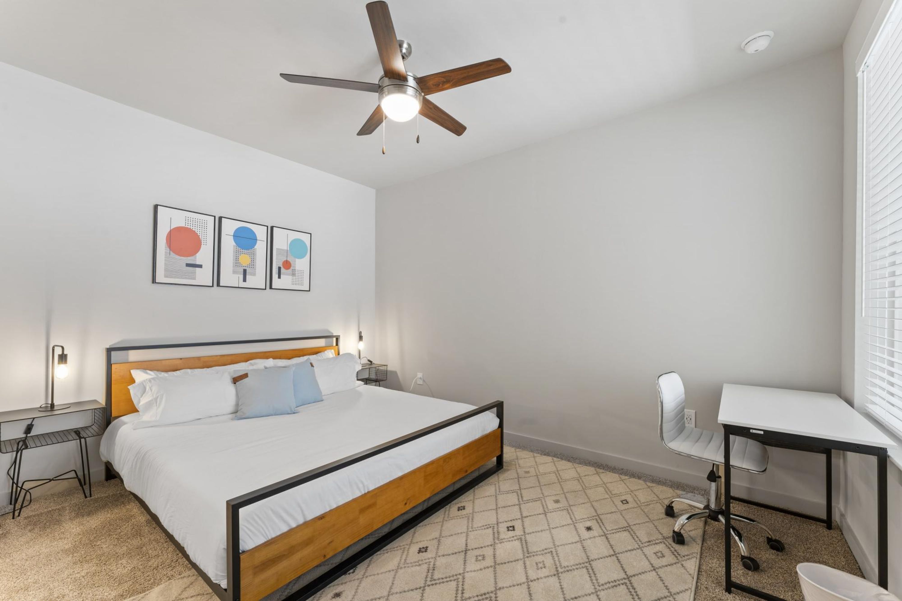 A bedroom with a bed, desk, chair, ceiling fan, and wall art.