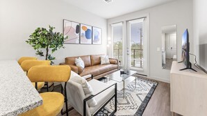A modern living room with a brown leather sofa, yellow chairs, a glass coffee table, and a large TV on a wooden cabinet. - Prime Soco Cozy 1BR King Suite w Pool Parking (Austin)