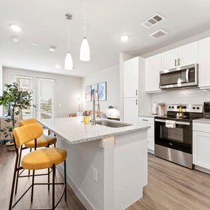 A modern kitchen with white cabinets, a granite island with bar stools, stainless steel appliances, and pendant lighting.