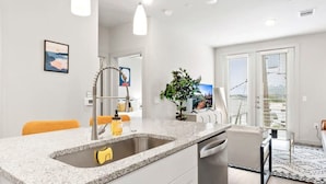 A modern kitchen with a granite countertop, stainless steel sink, and white cabinetry. There is a dining area with a table and chairs, and a large window with blinds. - Prime Soco Quaint 1BR King Suite w Pool Parking (Austin)