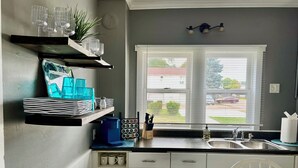 Una cocina con armarios blancos, una ventana con persianas, un lavamanos y una encimera con diversos objetos.
