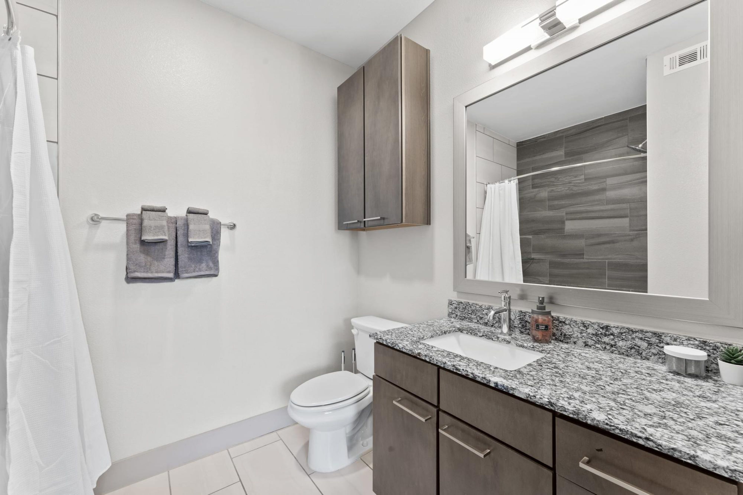 Un baño moderno con inodoro blanco, lavabo con encimera de granito y un espejo grande.
