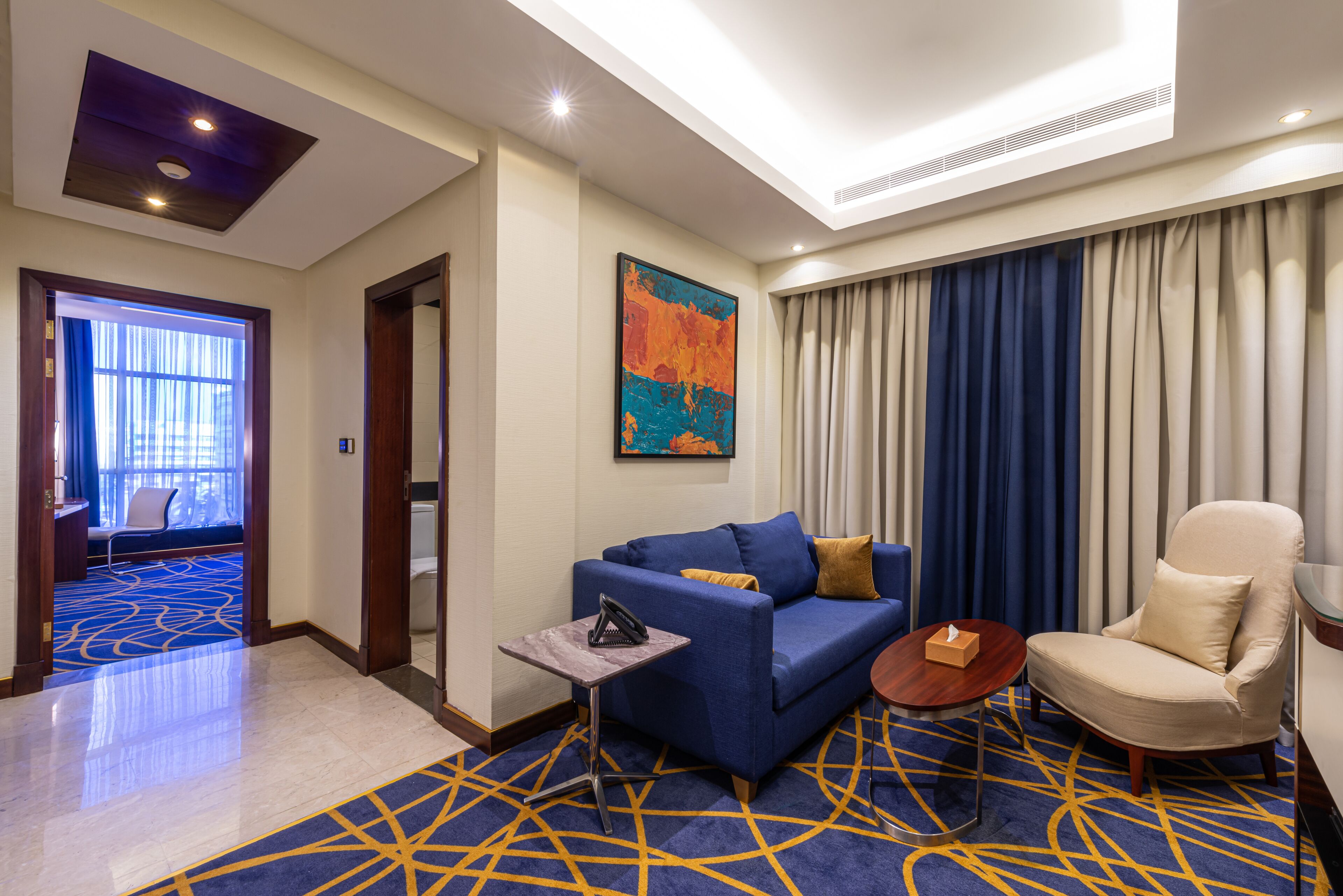 A hotel room with a blue sofa, a beige armchair, a small table, and a painting on the wall.