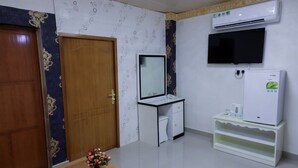 A room with a wooden door, a white desk with a mirror, a white refrigerator, and an air conditioning unit.
