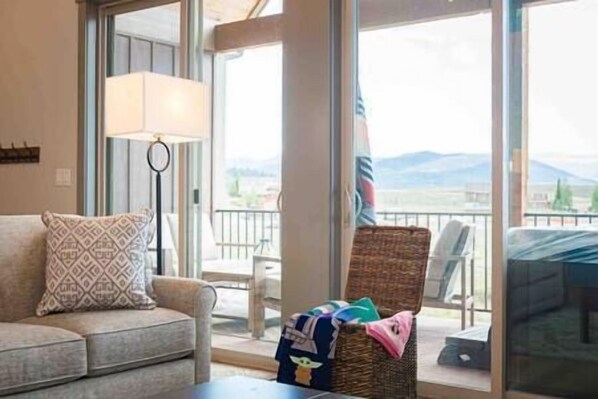 A living room with a sofa, a lamp, a wicker chair, and a view of mountains through large windows.