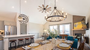 A modern kitchen with a central island, bar stools, a dining area set for six with a striped tablecloth, and a large clock on the wall. - New Cool 4BR Mountain Home: Views, Games, Hot Tub (Granby)