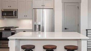A kitchen with white cabinets, a stainless steel refrigerator, a microwave, and a stove. There are three stools at the island. - New Cool 4BR Mountain Home: Views, Games, Hot Tub (Granby)
