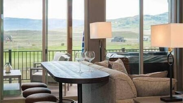 A room with a large window offering a view of rolling hills, a sofa, a table with glasses, and a lamp. - New Cool 4BR Mountain Home: Views, Games, Hot Tub (Granby)