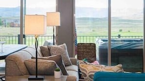 A room with a sofa, a table, and a view of a green landscape through large windows. - New Cool 4BR Mountain Home: Views, Games, Hot Tub (Granby)