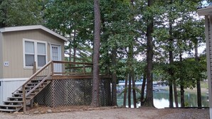 Une cabane dotée d’une terrasse en bois et d’un escalier menant à une zone abritée, le tout entouré d’arbres et bordé par un lac.