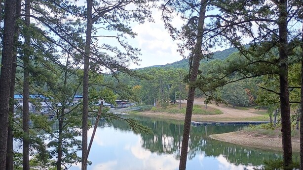 Une terrasse en bois avec une rambarde en métal, donnant sur un lac paisible, entourée d’arbres et offrant une vue lointaine sur un bâtiment.