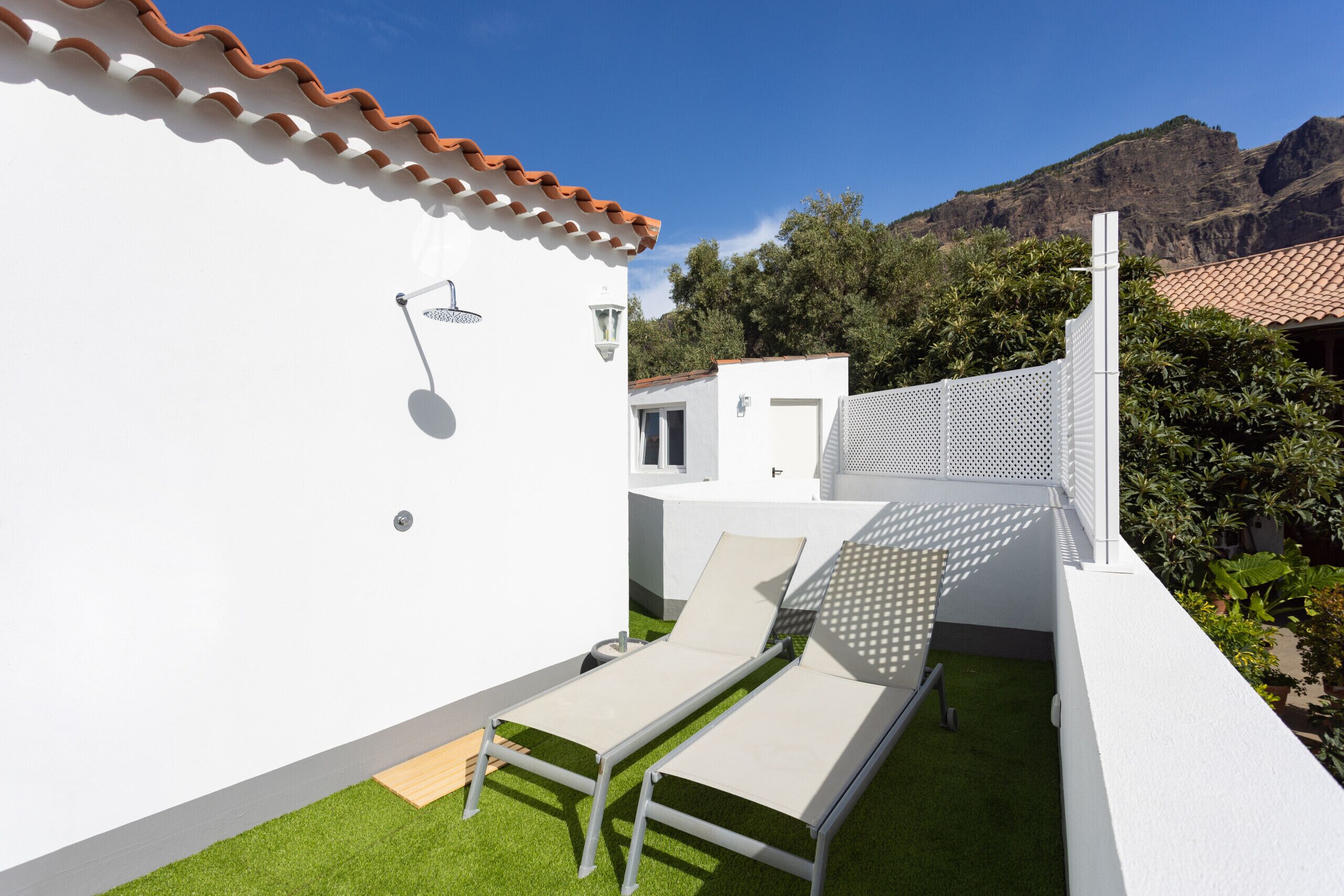 Ein moderner Außenbereich mit Sitzgelegenheit, einer weißen Wand, einem kleinen Garten und Bergblick.