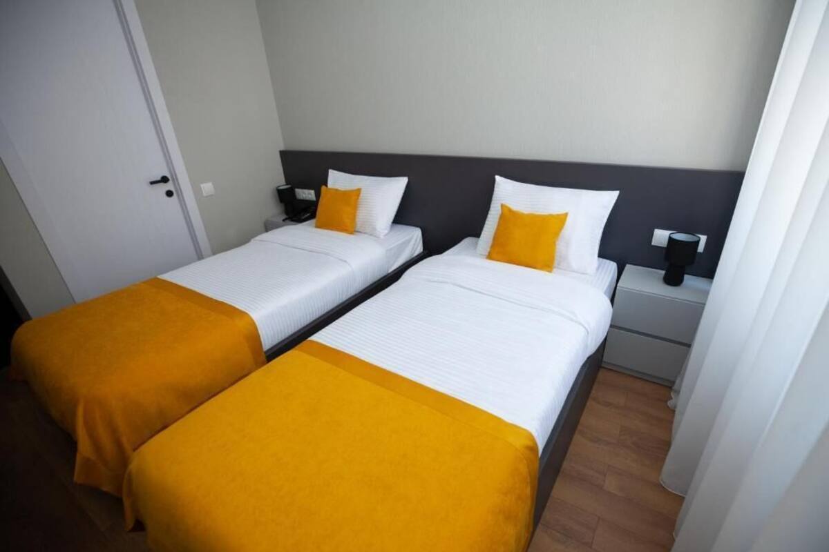 Two single beds with white bedding and yellow accents, a bedside table with a lamp, and a door in the background.