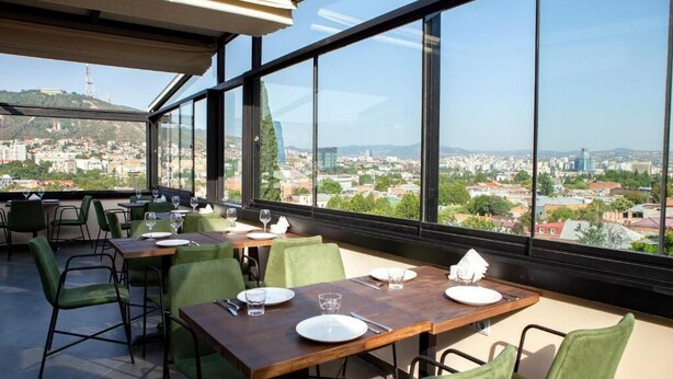 A restaurant with large windows offering a city view. Tables are set with plates, glasses, and cutlery. - Old City Boutique Hotel (Tbilisi)
