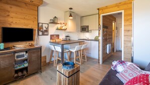 A compact kitchen with a dining area, wooden walls, a flat-screen TV, and a cozy seating area with a blanket. - Résidence Le Necou - Les Ménuires (Les Belleville)