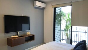 Une chambre moderne avec un grand lit, un téléviseur à écran plat fixé au mur, une petite console en bois et une porte-fenêtre coulissante donnant sur un balcon avec vue sur les palmiers.