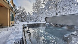 Een hottub met een glazen deksel, omringd door met sneeuw bedekte bomen en een gebouw met een beige gevel.