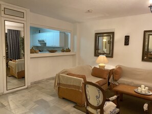 A living room with a sofa, a chair, a small table with candles, a lamp, and a kitchen area with cabinets and a window.