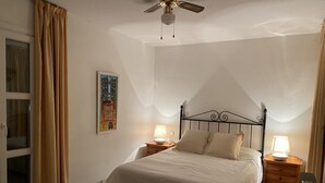 Une chambre à coucher avec un lit, deux tables de chevet avec des lampes, un ventilateur de plafond, une fenêtre avec des rideaux et un tableau au mur.