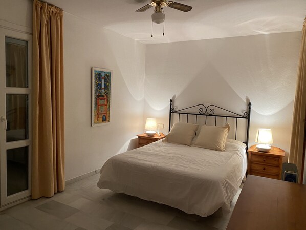 A bedroom with a bed, two bedside tables with lamps, a ceiling fan, a window with curtains, and a painting on the wall.