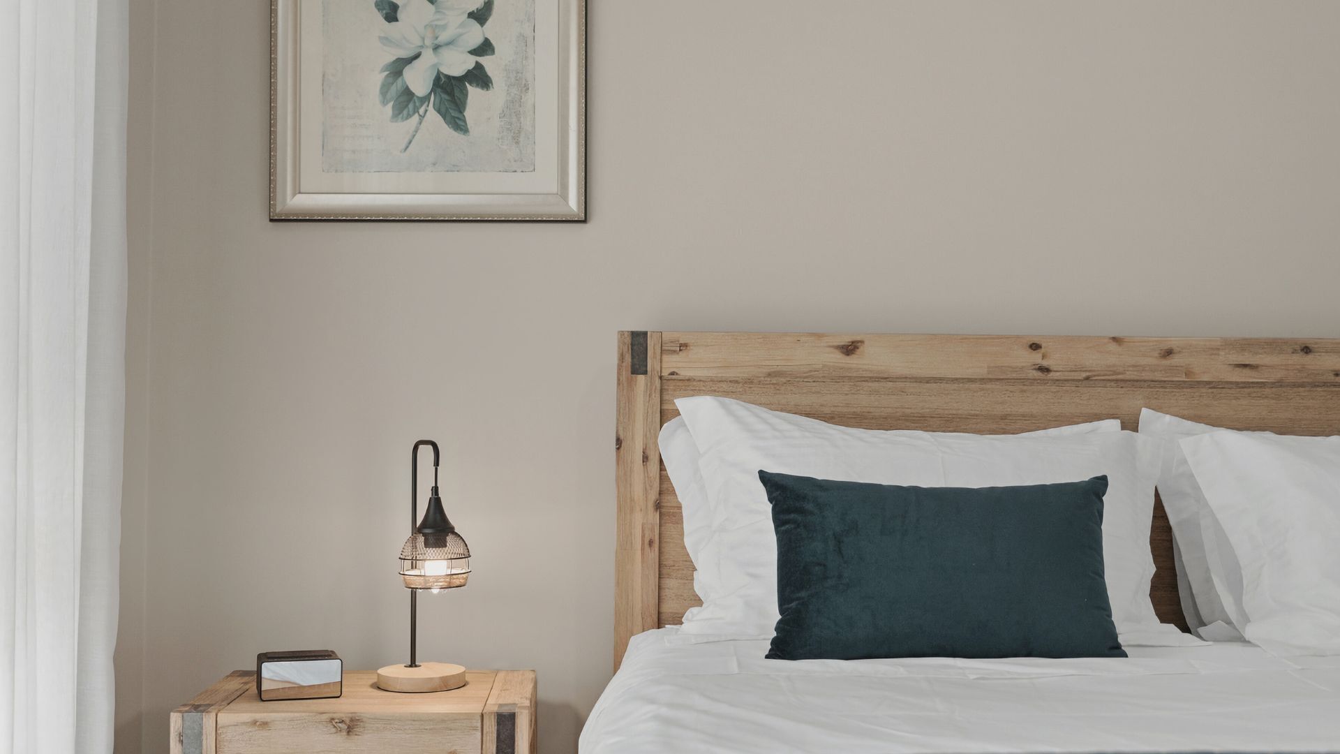 En pænt redt seng med hvide og mørkegrønne puder, et træsengegærde, et natbord med en lampe og et indrammet billede på væggen.