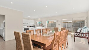 Un vaste espace de salle à manger avec une table et des chaises en bois, une cuisine en arrière-plan et une vue sur le jardin à travers de grandes fenêtres.