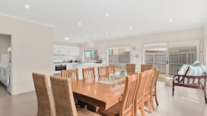 A spacious dining area with a wooden table and chairs, a kitchen in the background, and a view of the backyard through large windows.