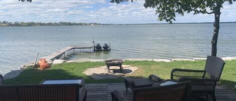Une terrasse dotée de meubles en osier, donnant sur un lac, un quai avec des bateaux et un foyer extérieur.