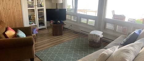 A living room with a sofa, armchair, TV, bookshelf, and a window with a view of the outdoors.