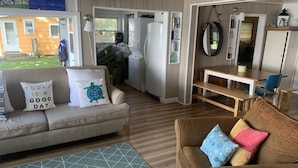 A cozy living room with a sofa, a brown armchair, and a coffee table. There is a dining area with a bench and chairs. A large window provides a view of the outside.