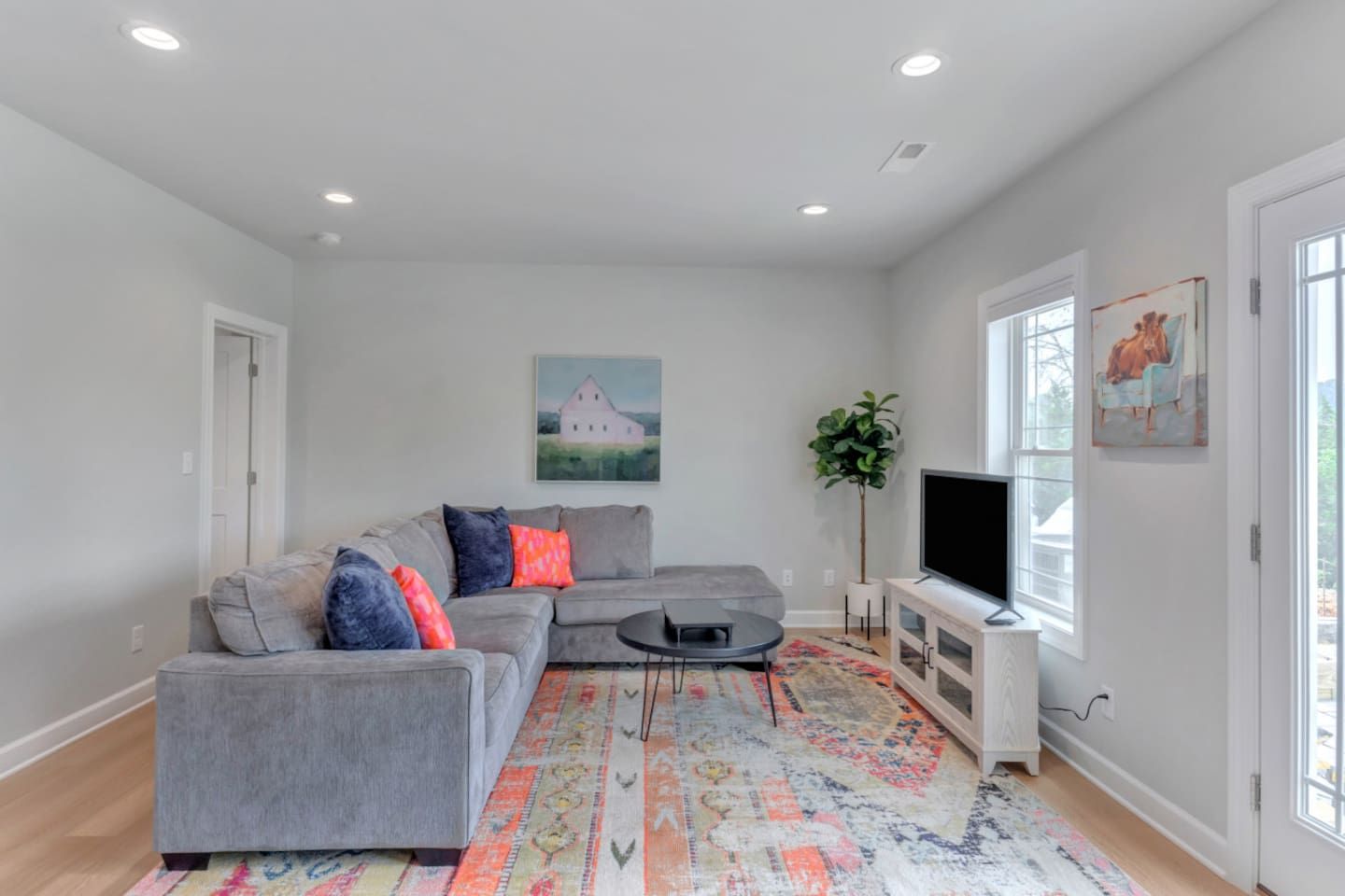 A living room with a grey sectional sofa, a flat-screen TV, a patterned rug, and artwork on the walls.