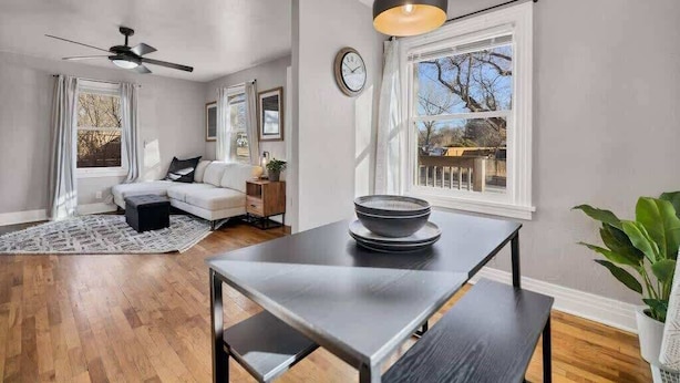 A modern living room with a dark wooden table, a ceiling fan, and a large window with a view of the outdoors. - Casa Mosaic! Downtown w BBQ & Workspace! (Colorado Springs)