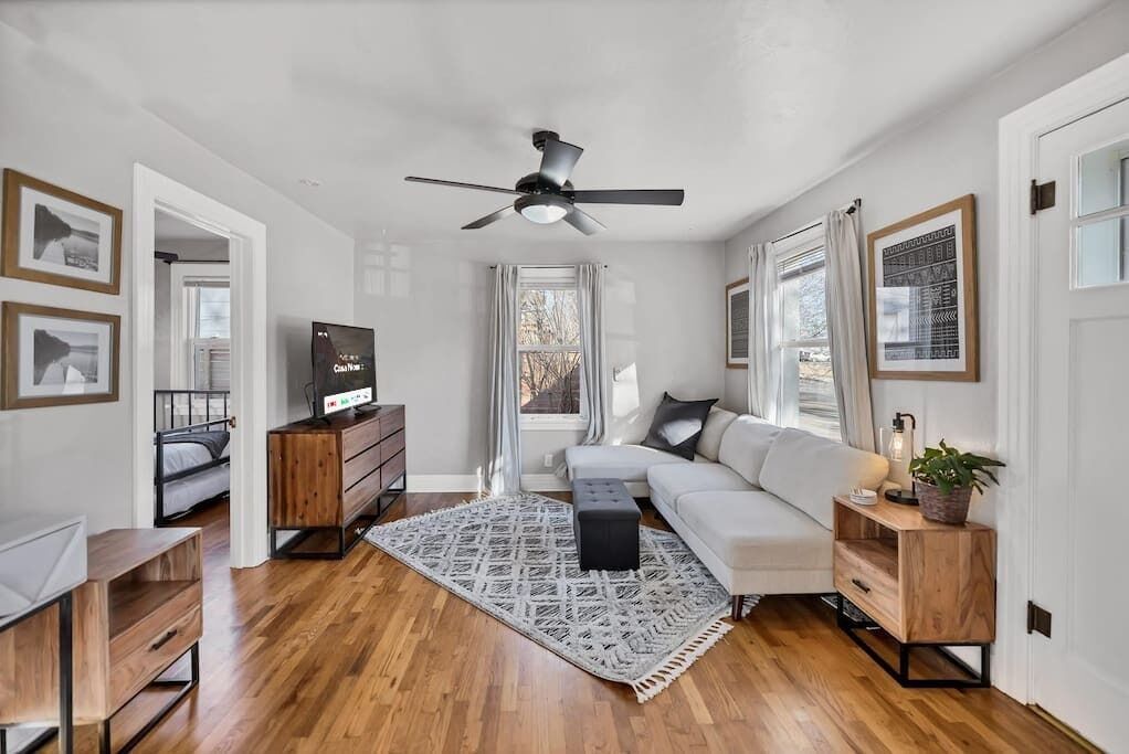 A modern living room with hardwood flooring, a white sofa, a black ottoman, a ceiling fan, and a large window with curtains.