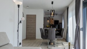 Ein modernes Wohnzimmer mit einer Couch, einem Essbereich mit Tisch und Stühlen, einer Küche im Hintergrund und einem großen Fenster mit Vorhängen.