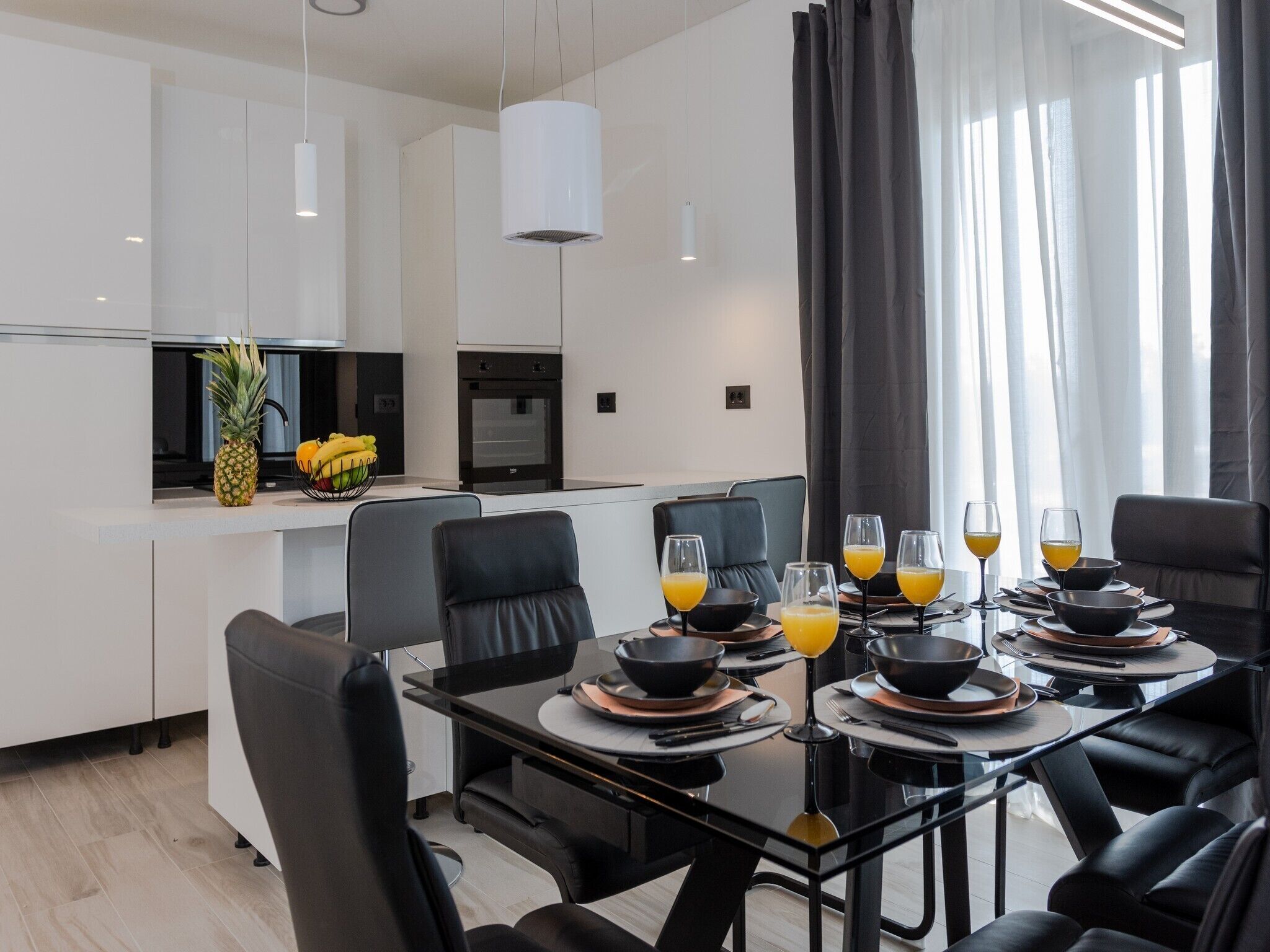 A modern dining area with a glass table, black chairs, and a kitchen in the background.